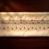 A neatly displayed white Parsi toran with a green vine and multi-colored floral embroidery pattern, finished with delicate white beadwork scallops and red bead accents.