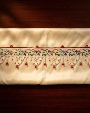 A neatly displayed white Parsi toran with a green vine and multi-colored floral embroidery pattern, finished with delicate white beadwork scallops and red bead accents.