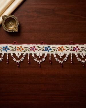 Top-down view of an intricate Parsi toran with multi-colored floral embroidery and white beaded scallops, styled on a dark wood table next to a brass diya and cream silk fabric.
