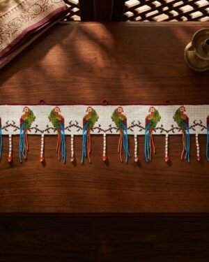 Top-down view of a traditional Parsi toran with intricate parrot embroidery and long blue and orange beaded tassels, resting on a dark wood surface next to a vintage brass lamp.