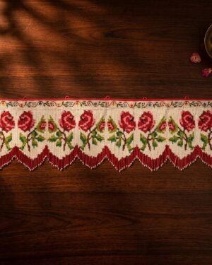Top-down view of a traditional Parsi toran with embroidered red roses and a red and white beaded fringe, displayed on a dark wooden table with scattered rose petals and a brass bowl.