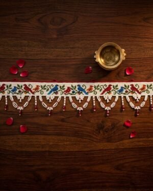 Top-down view of a white Parsi toran with detailed embroidery of colorful birds on a green vine. The piece is styled on a dark wood surface with red rose petals and a brass bowl.