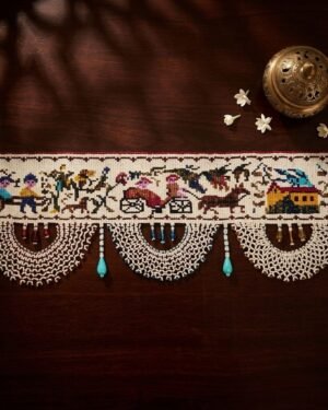 Top-down view of an intricate Parsi toran with embroidered scenes of people, horse-drawn carriages, and houses. It is finished with wide, semi-circular white beadwork and hanging turquoise beads, styled on a dark wood table with a brass incense burner.