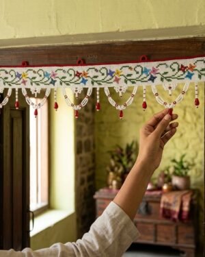 A person’s hand adjusts a traditional white Parsi toran hanging on a dark wood doorframe. The toran features vibrant floral embroidery and white beaded scallops with red droplets