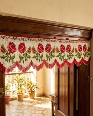 A decorative white Parsi toran hanging on a dark wood doorway, featuring a row of vibrant red roses with green leaves and a pointed fringe made of red beads.