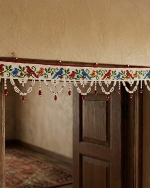 A traditional white Parsi toran hangs over a wooden doorframe, featuring a horizontal row of embroidered red, blue, and yellow birds with green vines, finished with white beaded scallops and red bead accents.