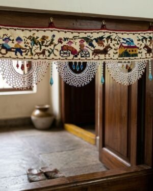A traditional white toran with multi-colored embroidery depicting village life hangs above a dark wood doorframe. The piece features large, elaborate white beaded scallops with blue accents, set against a bright home entryway with stone flooring.