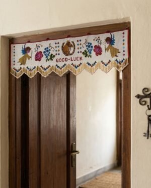 A decorative white Parsi toran hangs on a dark wood doorframe, featuring embroidered angels, floral patterns, and the words 'Good-Luck' with a gold beaded scalloped fringe. Vintage keys hang on the wall nearby.