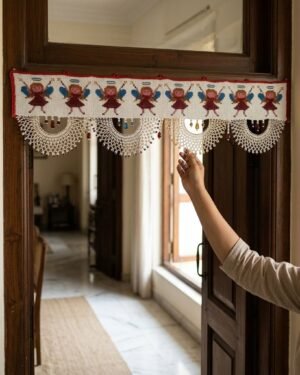A person’s hand touches the white beaded fringe of a Parsi toran hanging on a dark wood doorframe. The toran features a line of embroidered angels and hangs in a bright, traditional home interior.