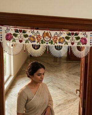A white Parsi toran featuring vibrant floral embroidery and a central horseshoe motif hangs on a dark wood doorframe. Below the toran, a woman in a light-colored saree is visible in the sunlit entryway of a traditional home.