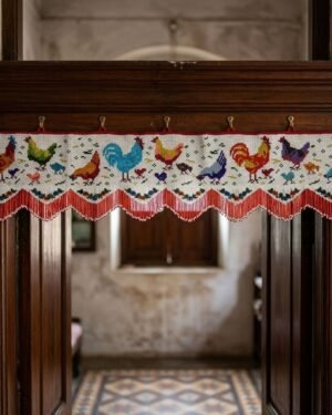 A white Parsi toran featuring a row of multi-colored embroidered roosters and chickens hangs on a dark wood doorframe, set against the sunlit interior of a traditional home with tiled floors.