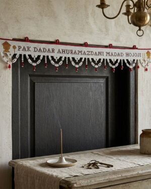 A traditional white Parsi toran hangs above a dark doorway, featuring the prayer 'PAK DADAR AHURAMAZDANI MADAD HOJOJI' embroidered in red. The piece is finished with white beaded scallops and red bead droplets, set in a rustic room with an incense stick and stone wall