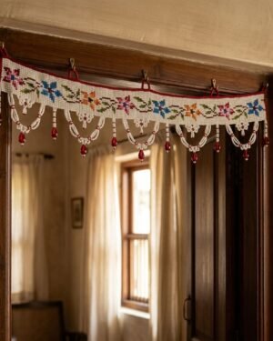 A white Parsi toran featuring multi-colored floral embroidery and white beaded scallops with red droplets, hanging on a dark wood doorframe in a traditional home interior.