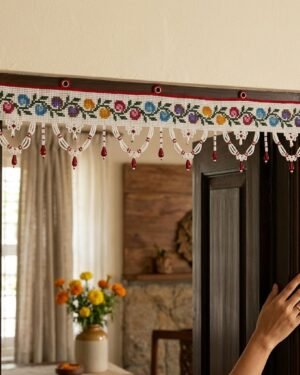 A handcrafted white Parsi toran featuring multi-colored floral embroidery and intricate white beaded scallops with red droplets. It is hanging on a dark wood doorframe in a traditional home interior where a hand is visible touching the door.