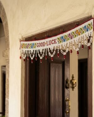 A white Parsi toran with 'GOOD LUCK' text and floral embroidery hangs on a dark wood doorframe, welcoming visitors into a traditional home entryway with arched stone architecture.