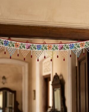 A white Parsi toran decorated with vibrant multi-colored floral embroidery hangs on a dark wooden doorway. Below the main band are delicate triangular beaded tassels in alternating colors, set against a blurred traditional home interior.