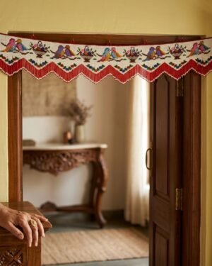A handcrafted white Parsi toran decorated with embroidered colorful birds and flower baskets hangs over a dark wooden doorframe. A person’s hand is visible in the foreground, and a sunlit room with traditional furniture is seen through the doorway.