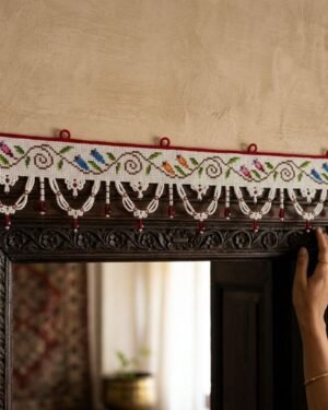 person’s hand reaches up to adjust a traditional Parsi toran hanging above a dark, carved wooden doorway. The white toran features a line of embroidered birds in various colors and is finished with intricate white beaded scallops accented with red droplets.