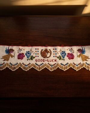 A horizontal view of a white Parsi toran featuring intricate embroidery of angels, roses, and a central 'Good-Luck' message, finished with a golden scalloped beaded fringe and resting on a dark wooden surface.