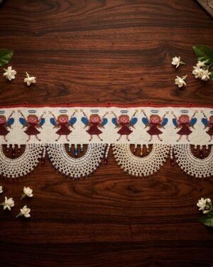Top-down view of a traditional Parsi toran featuring a series of embroidered angels with blue wings. The piece is finished with wide, intricate white beaded scallops and is styled on dark wood with scattered jasmine flowers.
