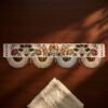 A flat view of an embroidered Parsi toran resting on a dark wood table. The design includes a horseshoe, flowers, and vines, finished with four large, semi-circular white beaded scallops with red bead accents