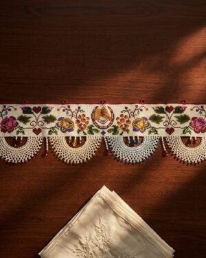 A flat view of an embroidered Parsi toran resting on a dark wood table. The design includes a horseshoe, flowers, and vines, finished with four large, semi-circular white beaded scallops with red bead accents