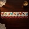 Top-down view of a handcrafted Parsi toran with vibrant rooster and hen embroidery. The piece is finished with a red and white pointed beaded fringe and is styled on a dark wooden table next to a brass oil lamp.