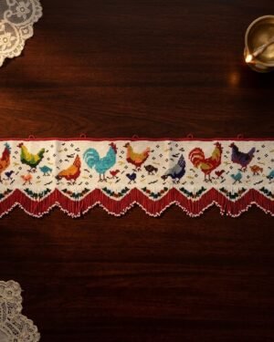 Top-down view of a handcrafted Parsi toran with vibrant rooster and hen embroidery. The piece is finished with a red and white pointed beaded fringe and is styled on a dark wooden table next to a brass oil lamp.