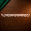 A flat view of a handcrafted Parsi toran resting on a dark wood surface. The white fabric is embroidered with a Zoroastrian prayer in red and accented by symbols of fire altars. It features an intricate white beaded fringe with hanging red beads.