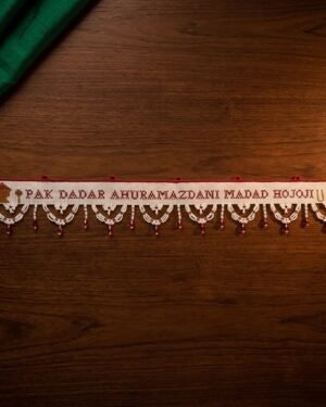 A flat view of a handcrafted Parsi toran resting on a dark wood surface. The white fabric is embroidered with a Zoroastrian prayer in red and accented by symbols of fire altars. It features an intricate white beaded fringe with hanging red beads.