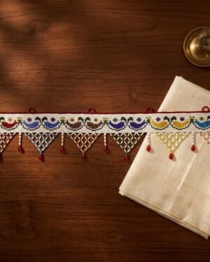 A flat view of a handcrafted Parsi toran resting on a dark wood surface. The design features colorful embroidered semi-circles and corresponding beaded lattice triangles, styled with a cream silk cloth and a vintage brass vessel.
