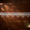 A flat, horizontal view of a white Parsi toran resting on a dark wood surface. The piece features a row of colorful embroidered flowers and a white scalloped beaded fringe, styled alongside a brass bowl.