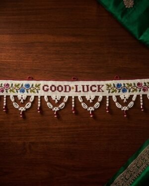 Top-down view of a traditional white Parsi toran featuring 'GOOD LUCK' embroidered in red, flanked by colorful floral motifs. The piece is finished with intricate white beaded scallops and red bead accents, styled on a dark wooden surface with green fabric.