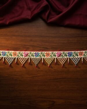A top-down view of a handcrafted Parsi toran resting on a dark wood surface with maroon fabric. The piece features a row of embroidered flowers and a fringe of colorful triangular beadwork lattice, showcasing intricate Parsi craftsmanship.