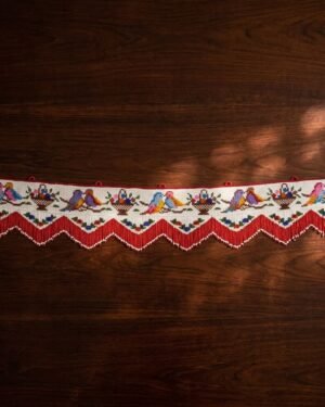 Top-down view of a traditional Parsi toran featuring intricate embroidery of birds in pairs and baskets of flowers. The piece is finished with a red and white pointed beaded fringe and rests on a dark wooden surface.