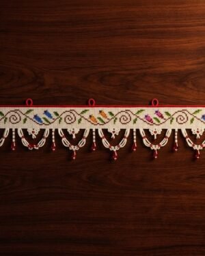 Top-down view of a white Parsi toran resting on a dark wood surface. The design features a continuous vine with pairs of multi-colored embroidered birds and a bottom fringe of white beaded scallops with hanging red beads.