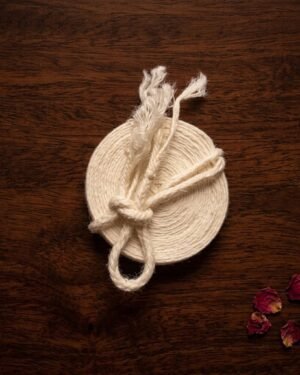 Top-down view of a sacred Kusti cord coiled into a disc on a dark wood surface, featuring a knot and tasseled ends, accented by scattered red rose petals