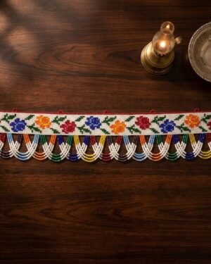 Top-down view of a handcrafted Moti no Toran with intricate beadwork depicting roses in various colors. The piece is styled on a dark wood table next to a lit brass oil lamp and a silver bowl.