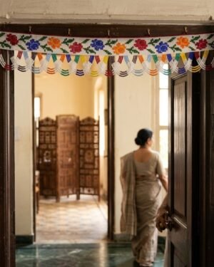 A traditional white beaded Parsi toran featuring multi-colored roses and a scalloped fringe hangs on a dark wood doorframe. A woman in a grey saree is seen from behind, walking through a sunlit traditional interior.