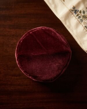 Top-down view of a maroon velvet Parsi topi on a dark wooden table next to an embroidered cloth.