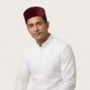 Studio portrait of a man wearing a structured maroon velvet Parsi cap and a crisp white formal tunic with a mandarin collar, set against an off-white background.