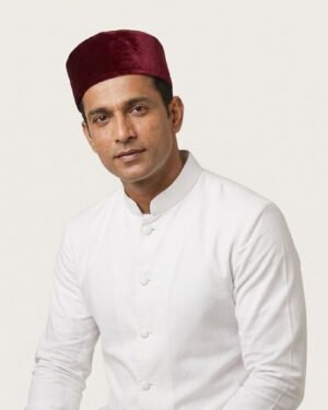 Studio portrait of a man wearing a structured maroon velvet Parsi cap and a crisp white formal tunic with a mandarin collar, set against an off-white background.