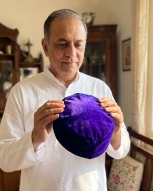 An older man wearing a white kurta looks down respectfully while holding a vibrant royal blue velvet Parsi cap in a traditional home interior.