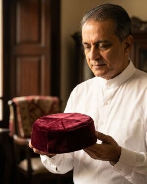 A man in a white formal shirt holding a maroon velvet Parsi topi with both hands.