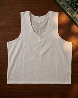 Top-down view of an unfolded white cotton Sudreh vest on a polished wood surface, accented by a green embroidered fabric in the corner.