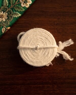 Top-down view of a coiled white wool Kusti on a dark wood table next to green floral embroidery.