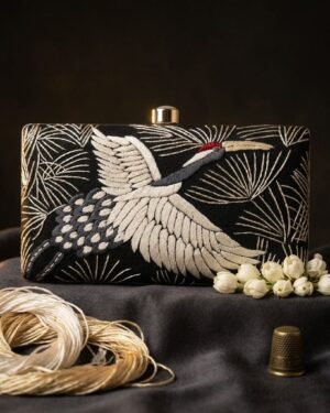 A professional product shot of a Parsi Gara embroidered clutch with a crane motif, styled with jasmine flowers, a spool of silk thread, and a thimble on a dark gray fabric