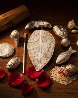 A collection of Parsi silver ceremonial items, including a silver fish, leaf, and betel nut, arranged on a wooden table with rose petals and jasmine flowers