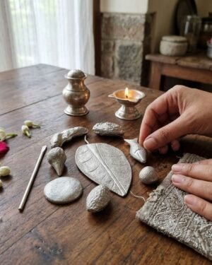 Close-up of hands carefully arranging silver Parsi ritual items on a wooden table next to a lit oil lamp and flowers.