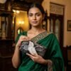 A woman in a green embroidered Gara sari holding a matching crane-motif clutch.